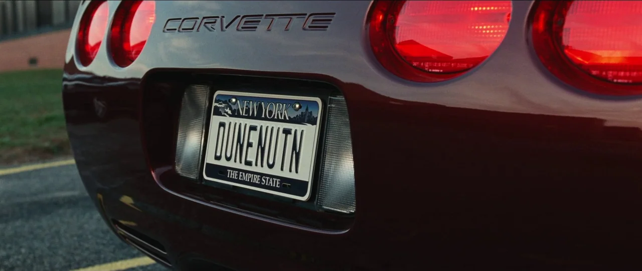 Bad Education (2019) movie still: closeup — The rear end of a dark red Chevrolet Corvette is positioned in a parking lot. A New York license plate…