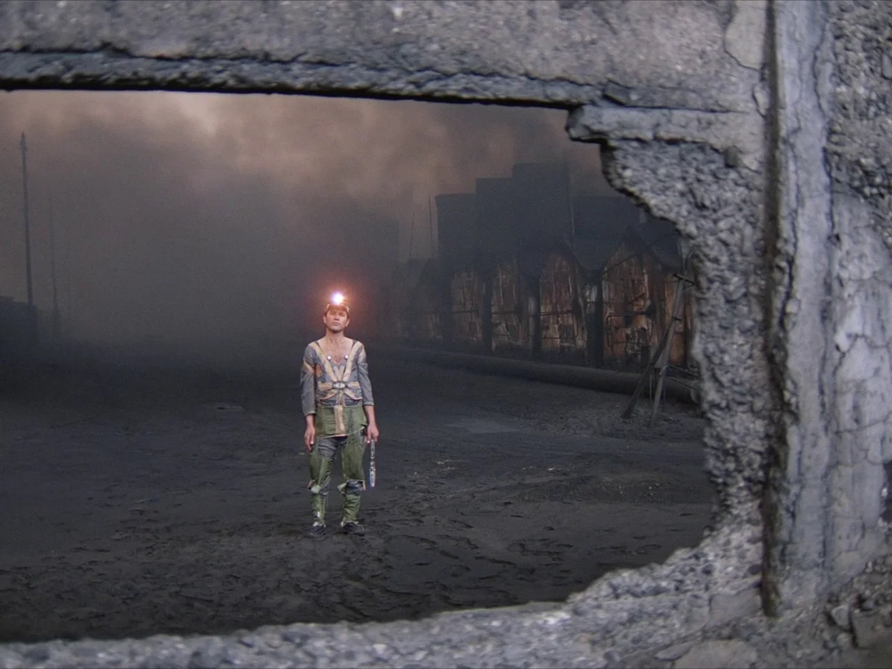 Kin-dza-dza! (1986) movie still: fullbody — A man stands in a desolate, smoke-filled industrial area with ruined brick buildings. He wears mining…