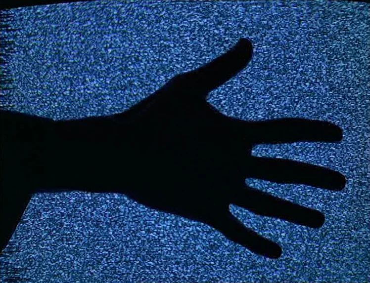 First Name: Carmen (1983) movie still: closeup — A silhouette of a human hand is superimposed over a blue television static background. The fingers…