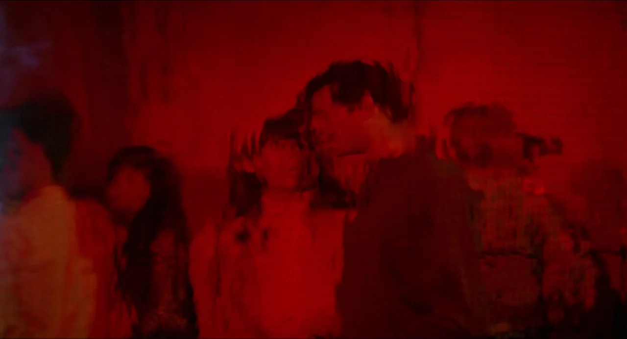 Medium Cool (1969) movie still: medium — A crowded nightclub interior features walls washed in intense, saturated red light. A young man whispers…