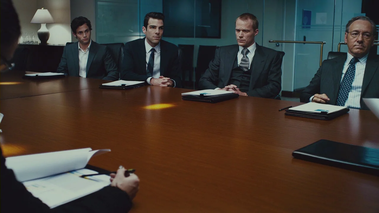 Margin Call (2011) movie still: wide — A sterile corporate boardroom features a long polished wooden table and glass wall partitions. Four men in…