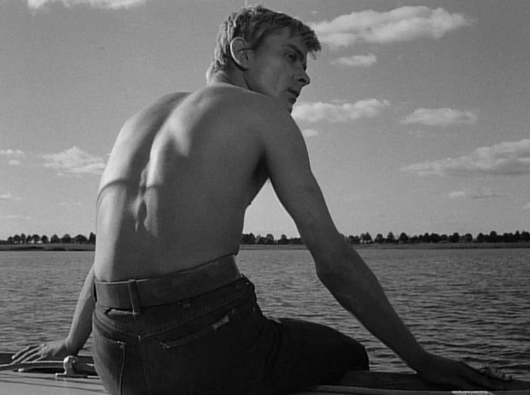 Knife In The Water (1962) movie still: medium — A man sits shirtless on the edge of a boat floating on a large lake. He turns his head to look back…