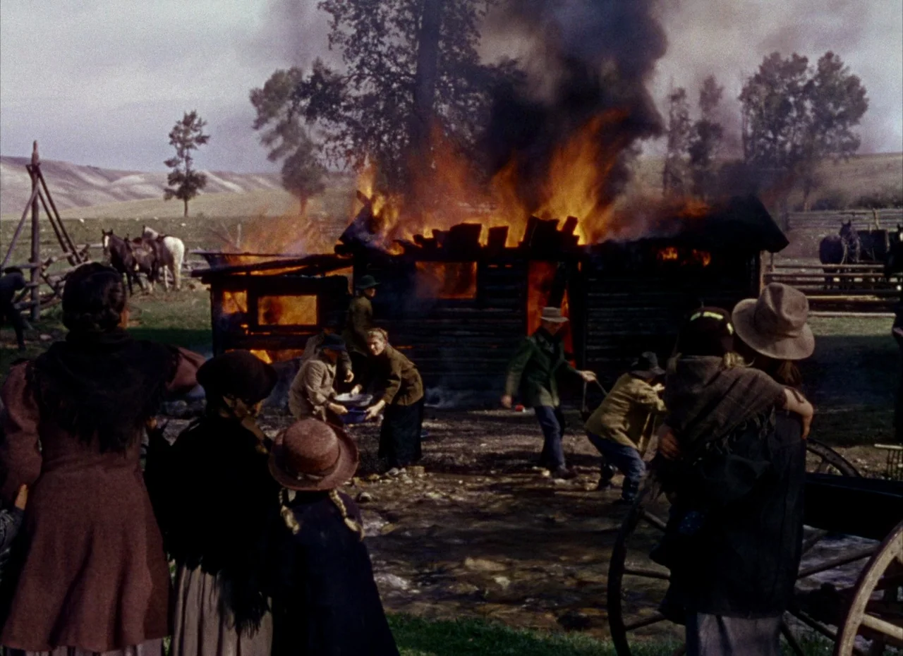 Shane (1953) movie still: wide — A rural homestead features a burning log structure surrounded by people in 19th-century attire. Several figures…