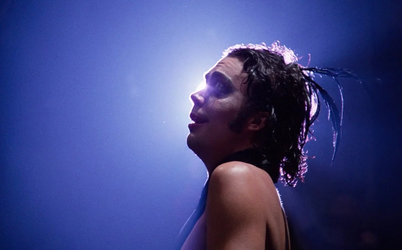 The Rocky Horror Picture Show (1975) movie still: closeup — A performer stands on a dark stage lit from behind by a harsh blue light. The individual…