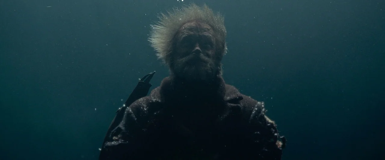 Sisu (2022) movie still: medium — A man is submerged in dark, deep water with particles suspended around him. He floats in place as his hair rises…