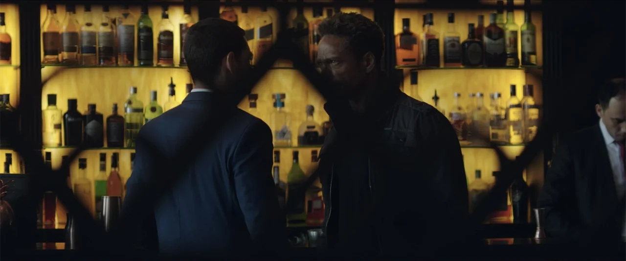 Redemption Day (2021) movie still: medium — A dimly lit lounge area features a long counter backed by illuminated glass shelves. Two men in dark…