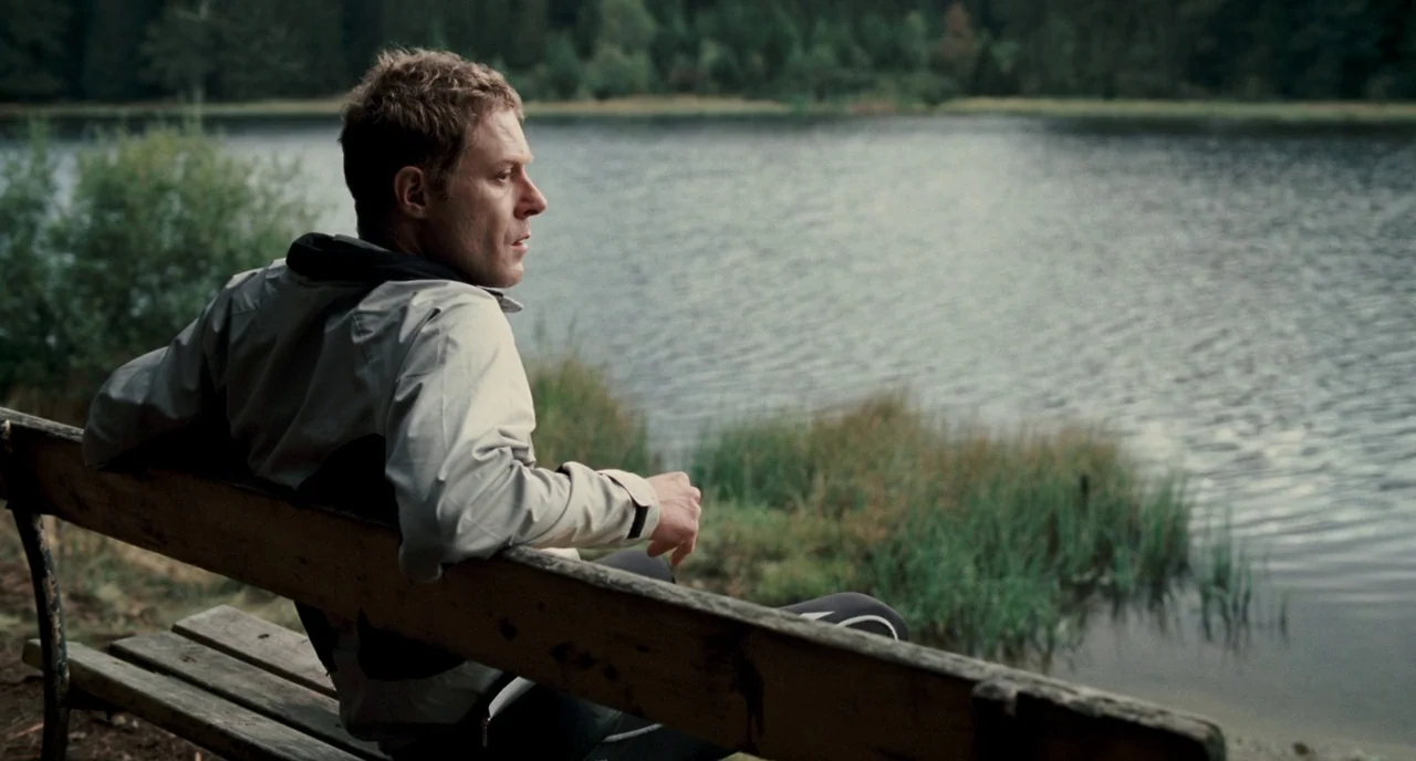 Revanche (2008) movie still: medium — A man sits on a wooden bench facing a calm body of water. Grassy reeds line the bank of the lake under an…