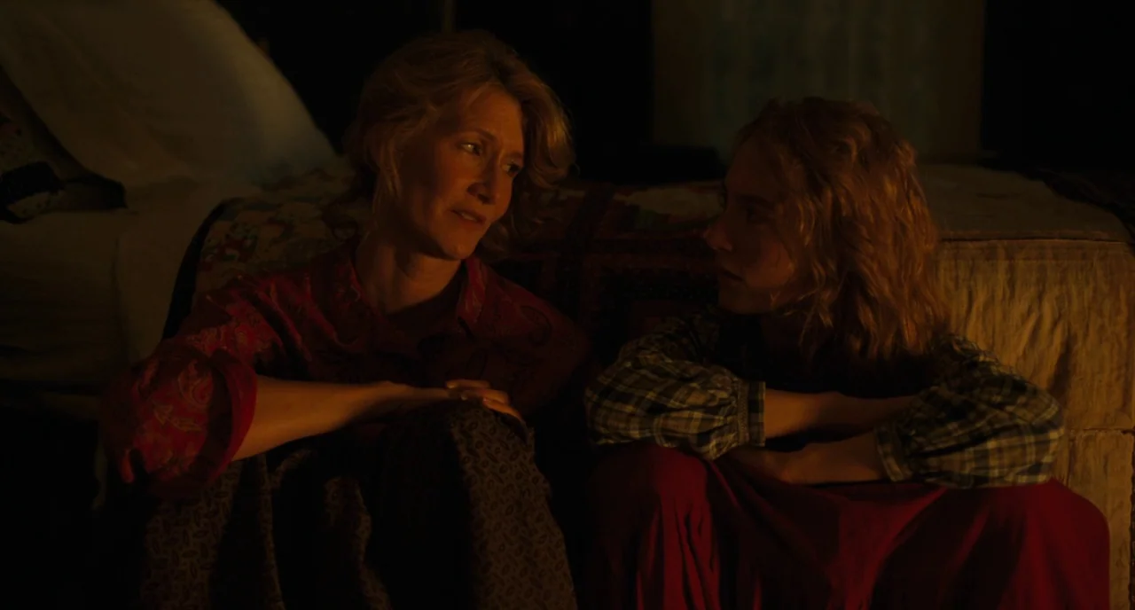 Little Women (2019) movie still: medium — A dimly lit room features a wooden bed with a quilt in the background. Two women sit on the floor facing…