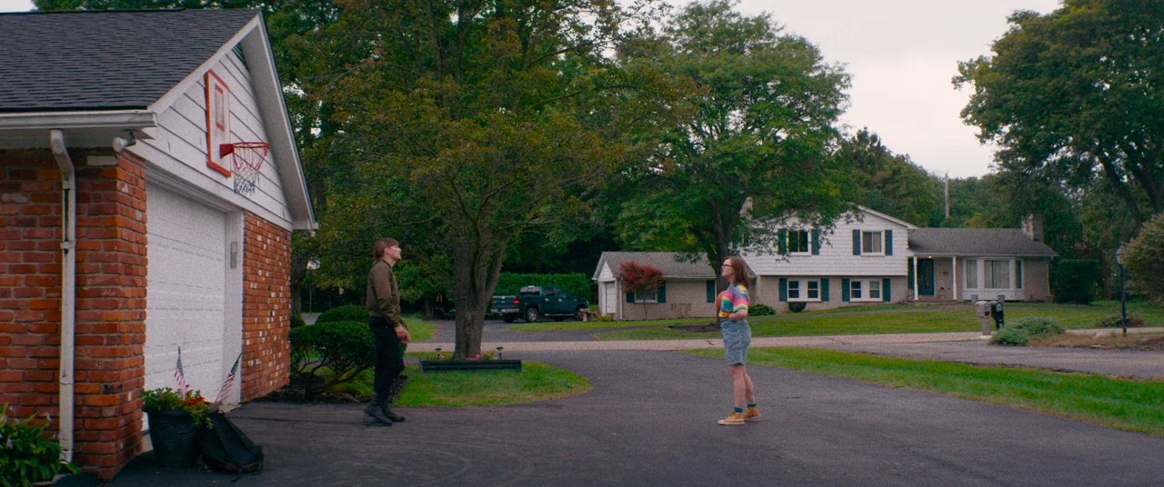 Dinner in America (2020) movie still: wide — A residential suburban driveway with a white garage on the left and houses beyond. Two young adults…
