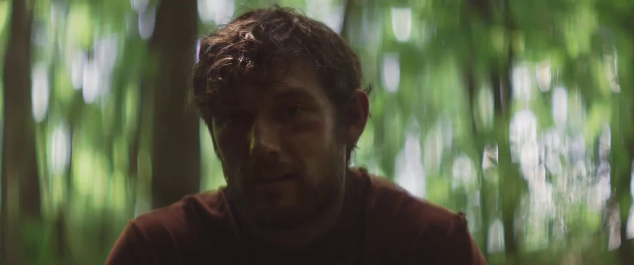 The Strange Ones (2017) movie still: closeup — A dense forest with out-of-focus foliage creates a soft green and brown background. A man sits in the…