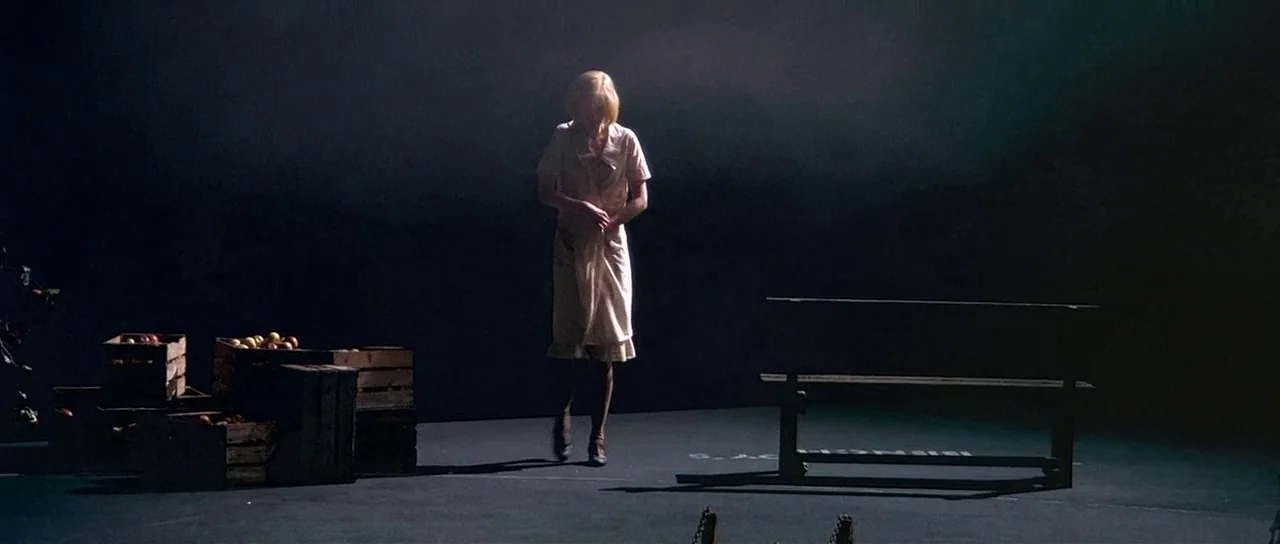 Dogville (2003) movie still: fullbody — A dark, minimalist stage set features only a wooden bench and stacked produce crates. A blonde woman in a…