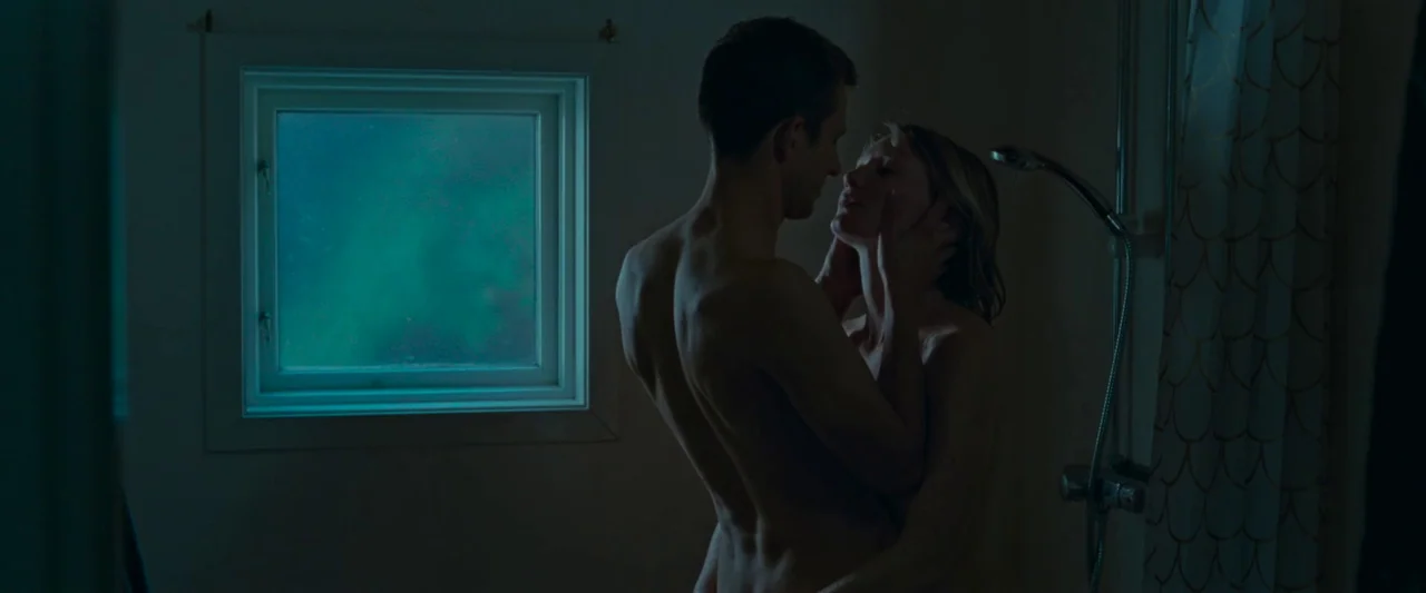 Bergman Island (2021) movie still: medium — A dark bathroom contains a shower stall with a single small window. A man and a woman stand together…