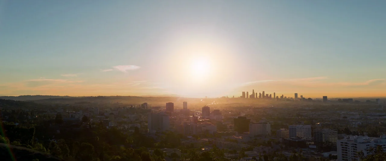 Captain Marvel (2019) movie still: establishing — A panoramic view of the Los Angeles skyline at golden hour with hazy air. The sun hangs low over…