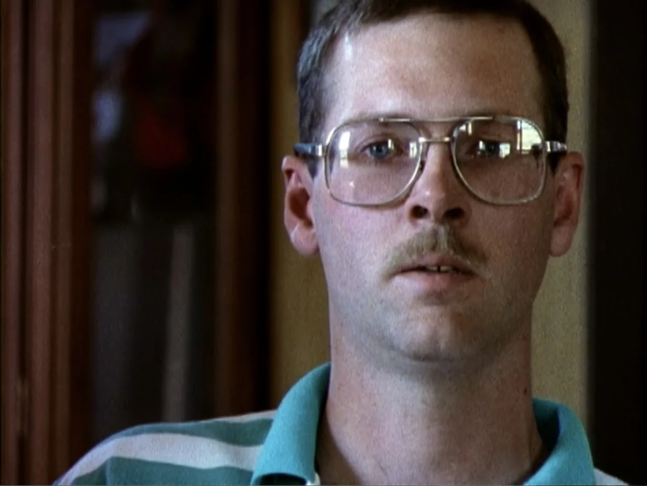 5 American Handguns - 5 American Kids (1995) movie still: closeup — A man in a polo shirt sits indoors during the daytime. The man stares forward…