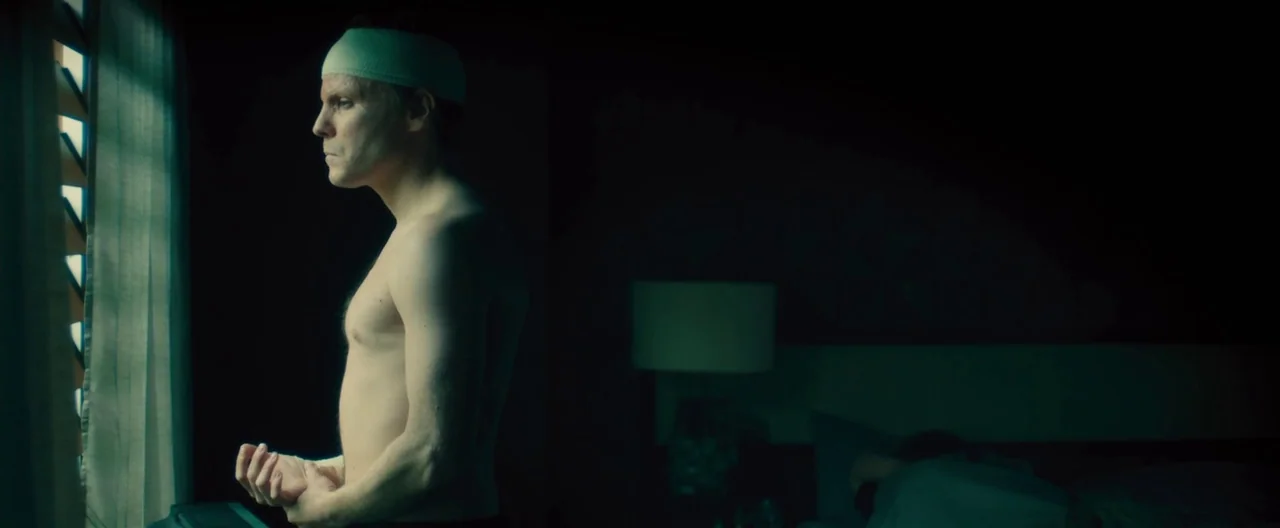 Rush (2013) movie still: medium — A dimly lit room with a man standing at a window with horizontal blinds. Another person lies on the bed in the…