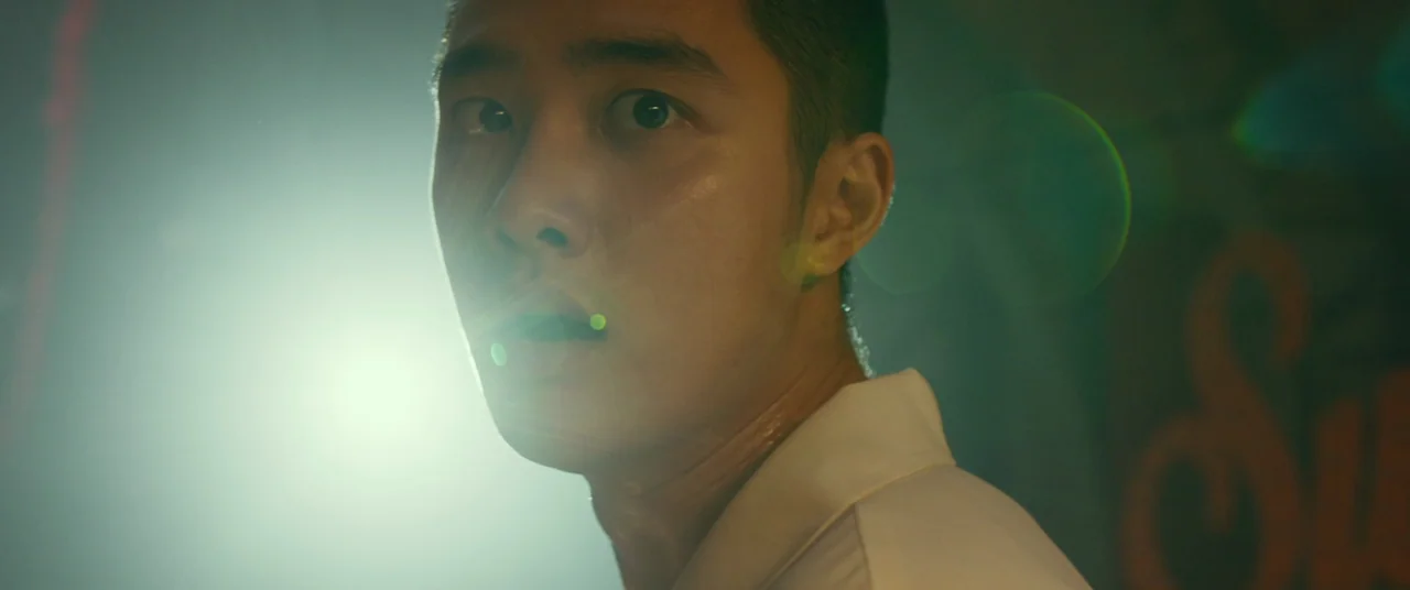 Swing Kids (2018) movie still: closeup — A dark outdoor space with indistinct textures visible in the background. A young man turns his head toward…