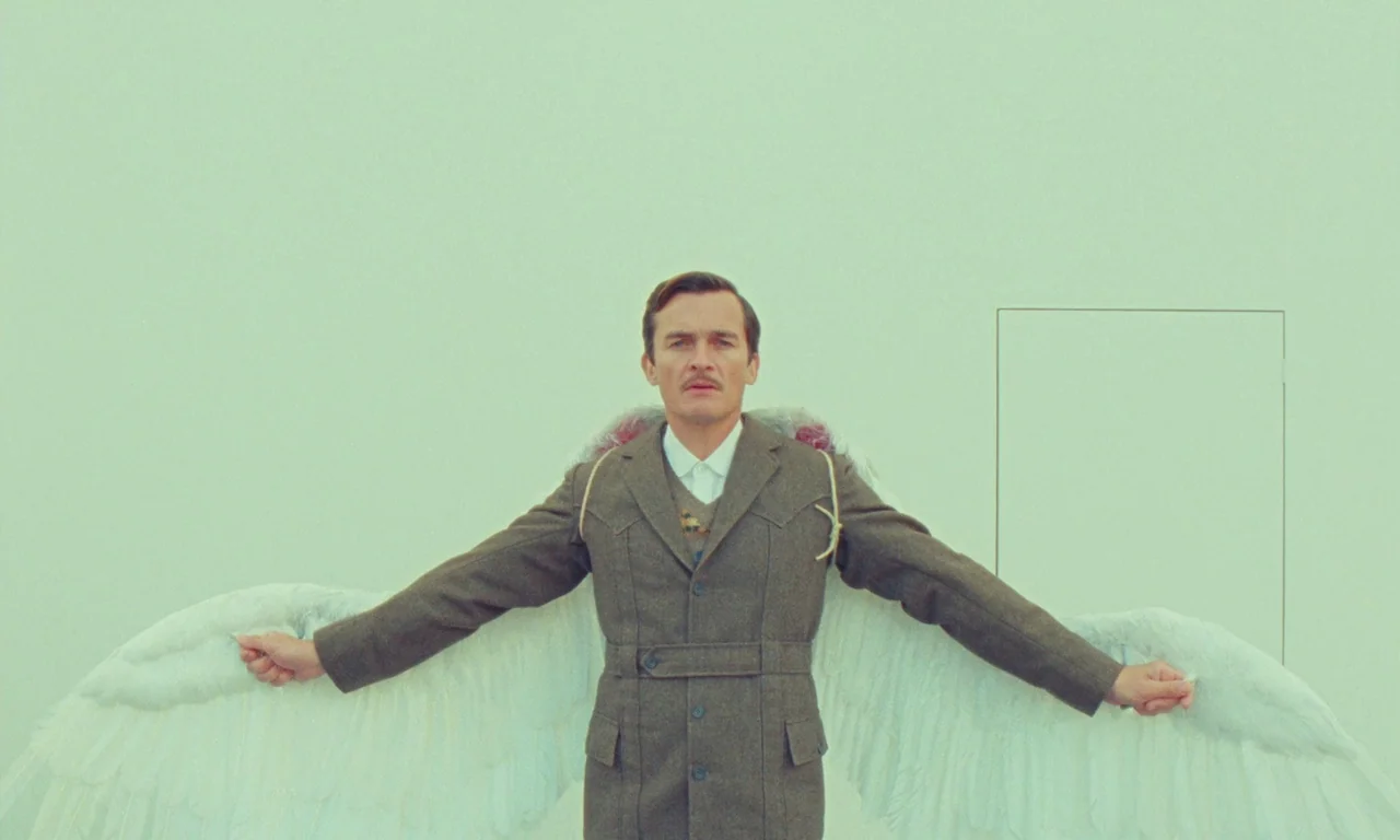 The Swan (2023) movie still: medium — A man stands centered against a flat, light-green wall with a rectangular door outline. He wears a brown…