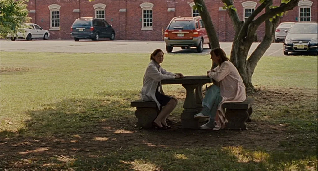 Choke (2008) movie still: wide — Two women are seated at a stone table in a grassy area before a building. The women talk while several parked cars…