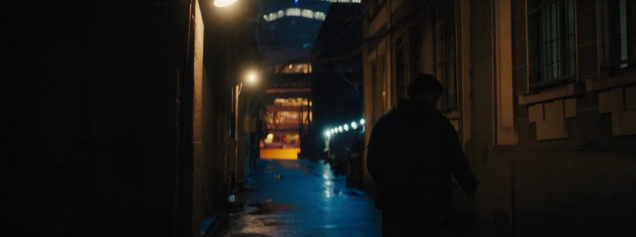 Dead Pigs (2018) movie still: medium — A narrow dark urban passage is illuminated by yellow overhead streetlights. A person walks away from the…