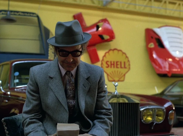 Keep Your Right Up (1987) movie still: medium — A man stands in front of a yellow wall adorned with automotive memorabilia. Vintage automobiles are…