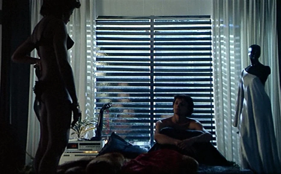 Diva (1981) movie still: fullbody — A dark bedroom interior features a large window covered by horizontal metal blinds. A woman stands in front of…