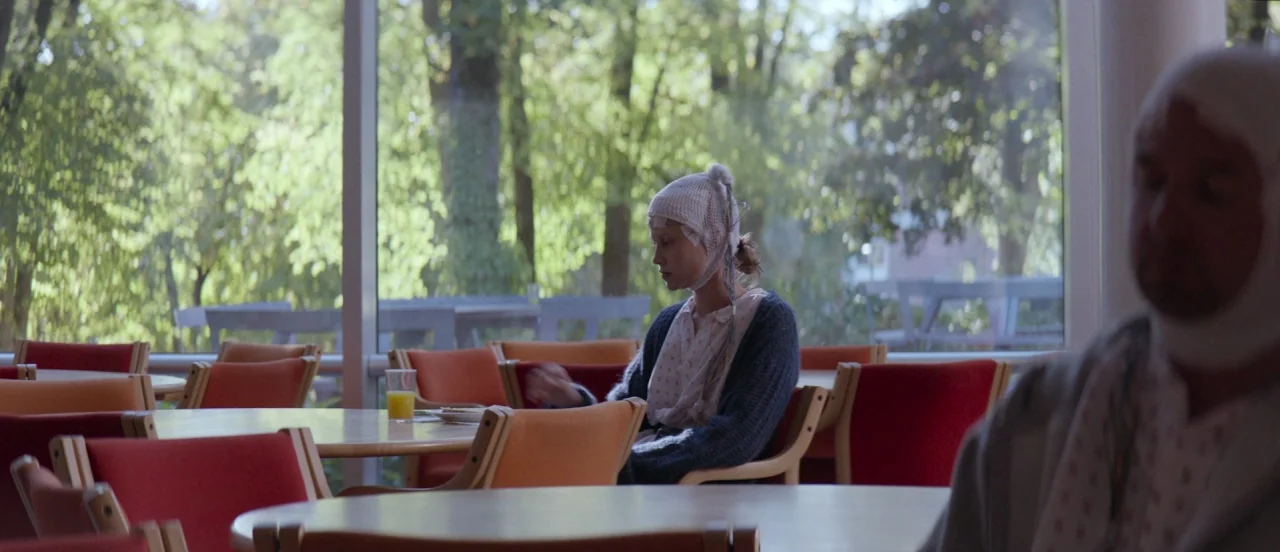 Thelma (2017) movie still: medium — A quiet, modern hospital cafeteria features orange chairs and tables by large windows. A woman wearing an EEG…