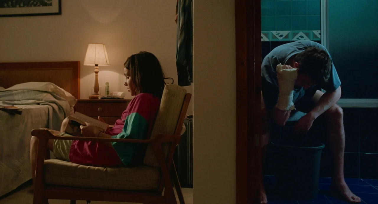 Aftersun (2022) movie still: wide — A split composition shows a young girl reading in a motel room bedroom area. To her right, a man with a bandaged…