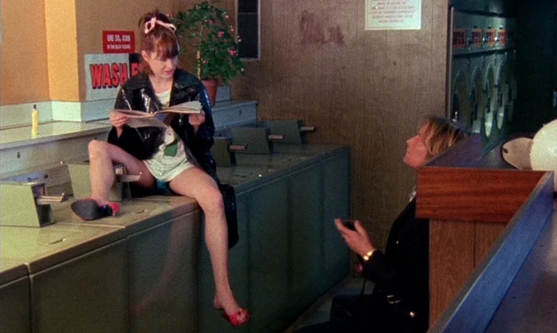 Jubilee (1978) movie still: medium — A 1980s laundromat features green industrial washing machines and faux-wood panel walls. A woman sits on a…