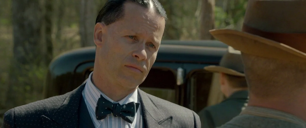 Lawless (2012) movie still: medium — A man in a formal 1930s suit stands in a sunny, wooded area. He faces another man while an antique car is…