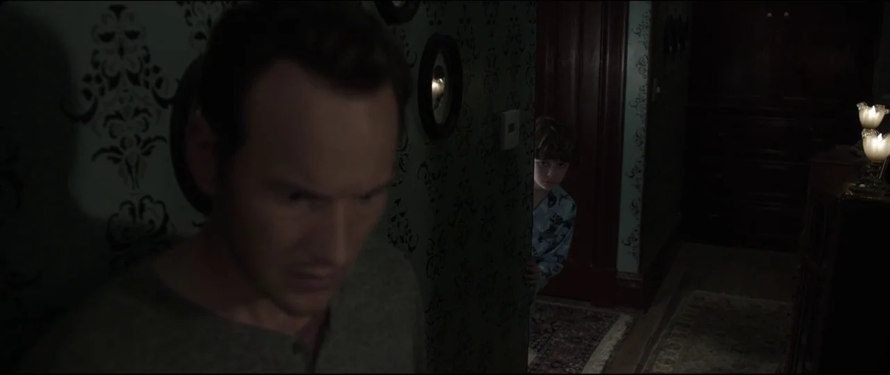 Insidious: Chapter 2 (2013) movie still: medium — A dimly lit hallway features dark patterned wallpaper and vintage wall decorations. A man walks in…