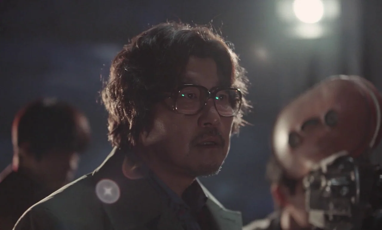 Cobweb (거미집) (2023) movie still: closeup — An interior production environment with a film crew in the low-lit background. A man wearing glasses…