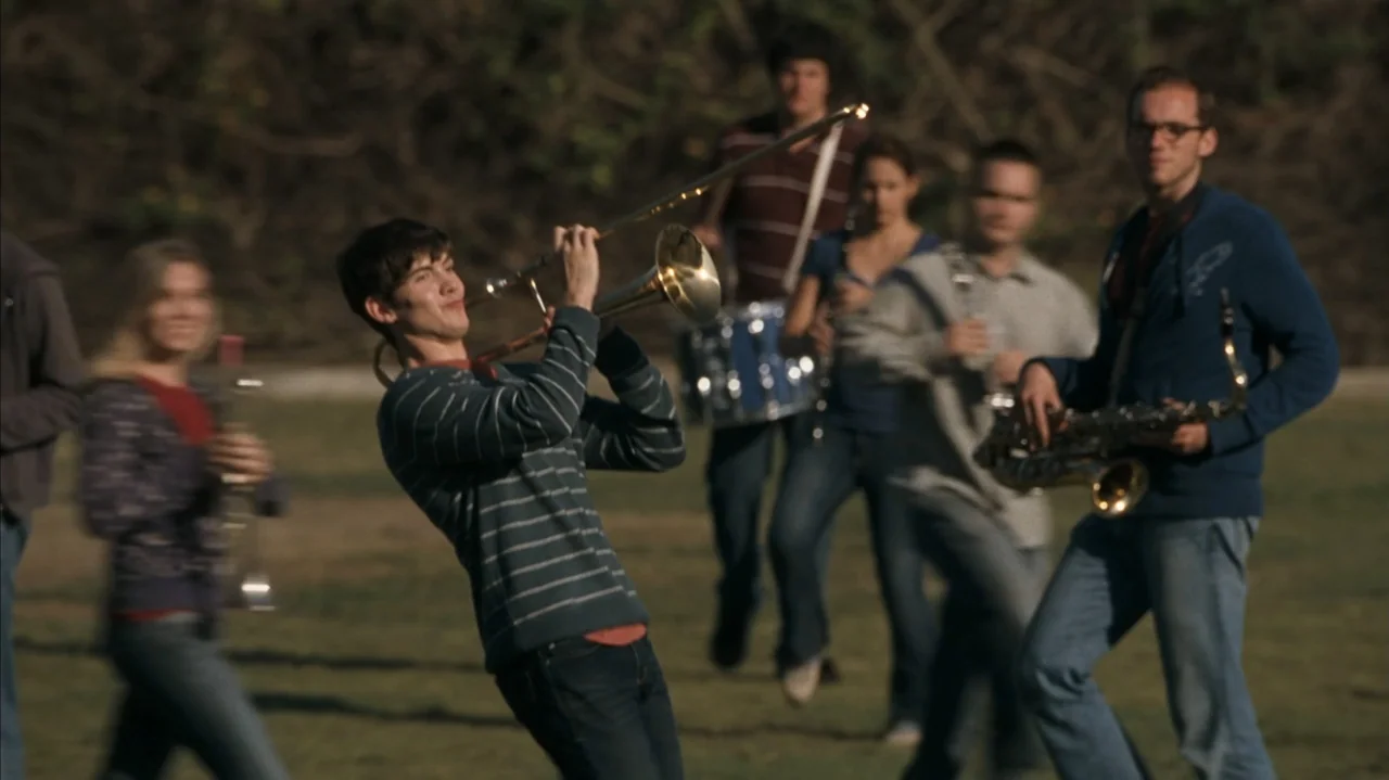F Valentine's Day (2026) movie still: medium — A group of musicians stands on a grassy field on a sunny day. A young man in a striped sweater plays…