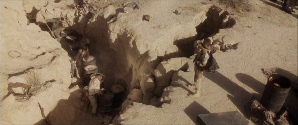 Three Kings (1999) movie still: wide — A deep rocky trench in a dry desert environment contains several iron barrels. Multiple workers move around…