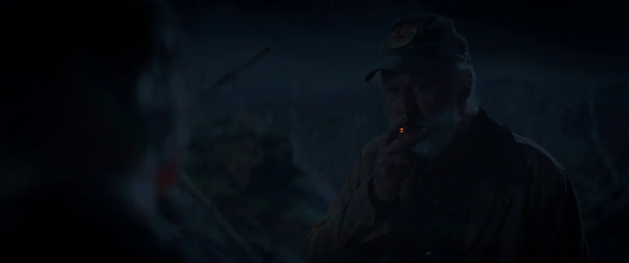Pet Sematary (2019) movie still: medium — An elderly man in a cap stands in a dark, open field at night. He holds a glowing cigarette to his mouth…