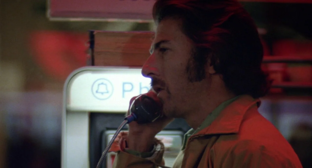 Straight Time (1978) movie still: medium — An outdoor night setting featuring a vintage public telephone kiosk with a glowing sign. A man stands in…