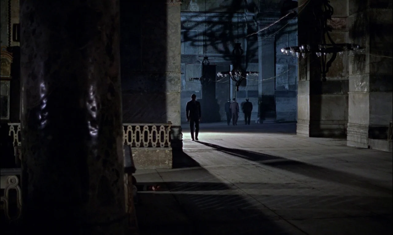 From Russia With Love (1963) movie still: wide — A dark stone interior features tall marble columns and hanging wrought iron chandeliers. A man in a…