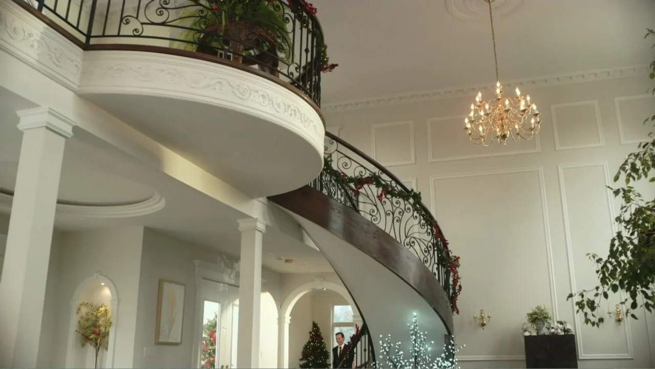 Driving Madeleine (2022) movie still: establishing — A grand, high-ceilinged mansion foyer features a large curved staircase with ornate iron…