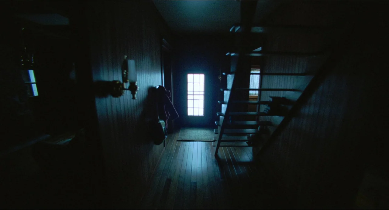 The Lodge (2019) movie still: establishing — A dark, narrow residential hallway features wood-paneled walls and a wooden staircase on the right. A…