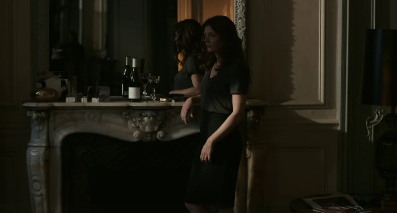 Bastards (2013) movie still: medium — A dimly lit room features a classical marble fireplace with an oversized mirror above. A woman stands in…