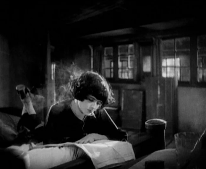 Sunrise (1927) movie still: medium — A dimly lit room contains a bed with a woman lying across it. She is smoking a cigarette while looking down at…