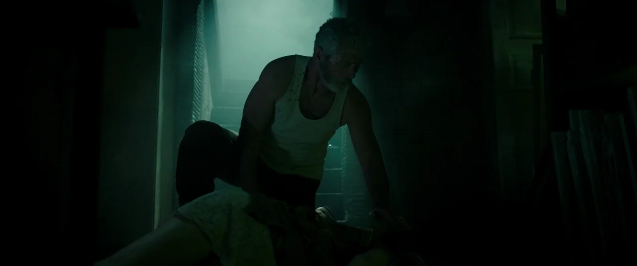 Don’t Breathe (2016) movie still: medium — A dimly lit basement features concrete walls and dusty atmosphere. An athletic man in a tank top kneels…