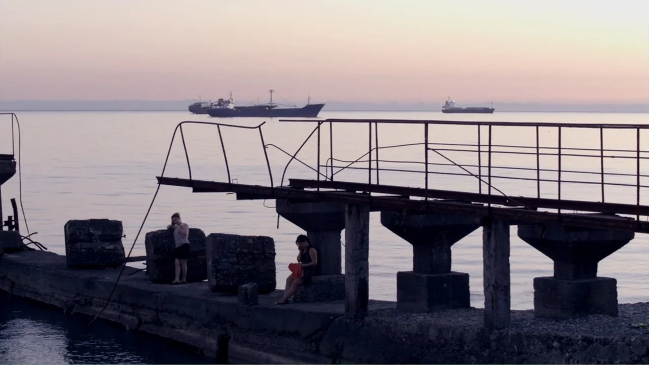 Letters to Max (2014) movie still: wide — A concrete pier extends into calm water under a pale twilight sky. Two women stand and sit on the pier…