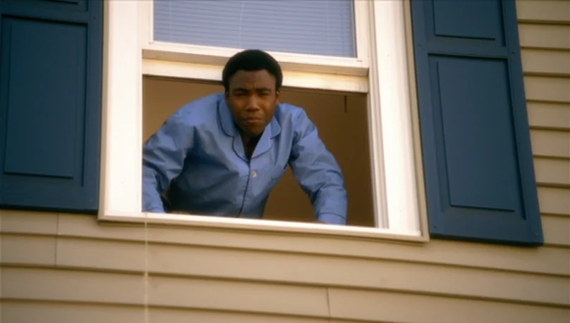 Mystery Team (2009) movie still: medium — A house exterior features tan horizontal siding with dark blue window shutters. A man leans forward from…