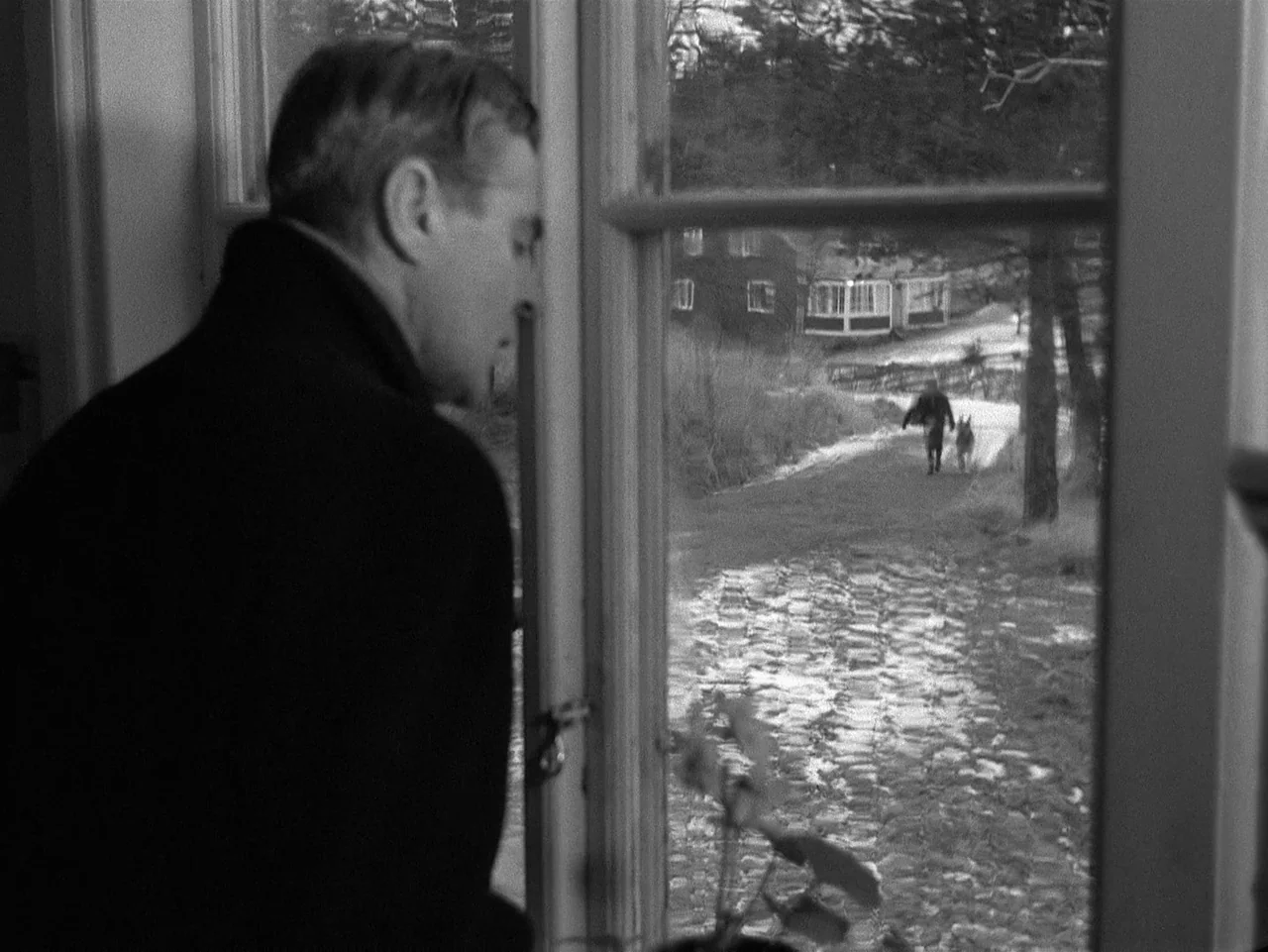 Winter Light (1963) movie still: medium — A man stands inside looking out a window at a cobblestone path. A distant figure walks their dog along the…