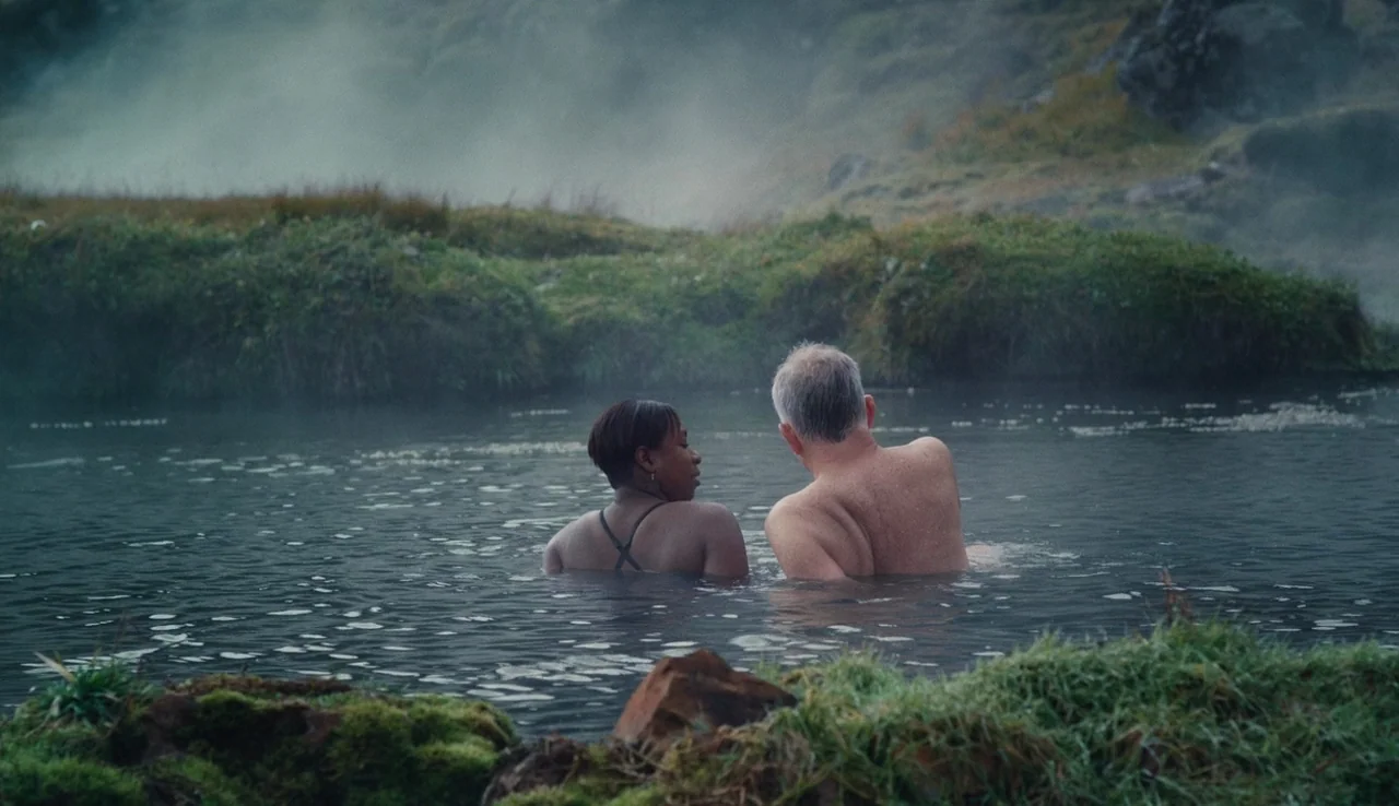 Land Ho! (2014) movie still: wide — A natural geothermal pool is set against a mossy volcanic landscape with rising steam. A man and a woman are…