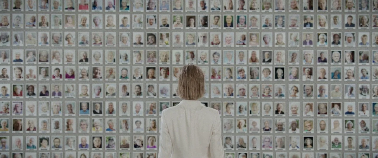 I Care A Lot (2020) movie still: medium — A large grid of hundreds of small portrait photographs is arranged on a wall. A woman in a light-colored…