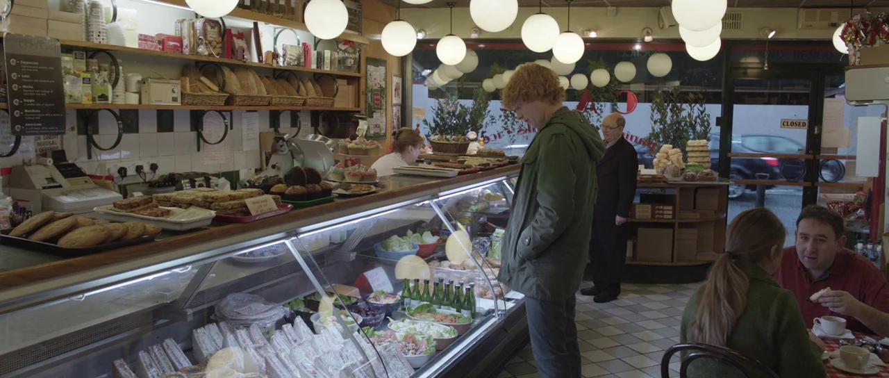 Frank (2014) movie still: wide — A food counter inside a small, brightly lit shop with glass cabinets of sandwiches. People stand and sit in the…