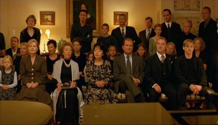 The Piano Teacher (2001) movie still: wide — A crowded room decorated with framed paintings contains multiple rows of seated people. The subjects…