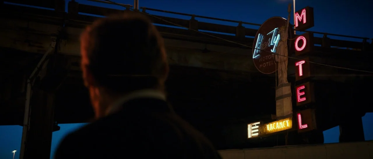 Logan (2017) movie still: medium — A man stands beneath a highway overpass at night near a glowing neon sign. The sign features a statue of the…