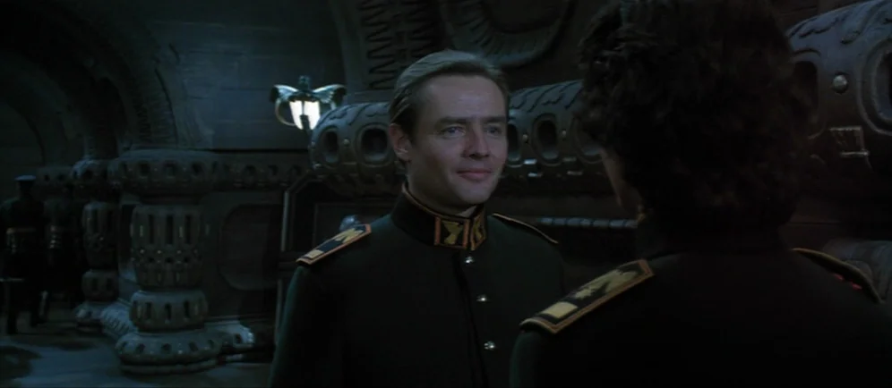 Dune (1984) movie still: medium — A dimly lit metallic hallway features ornate carved wall patterns and wall-mounted light fixtures. A man in a dark…